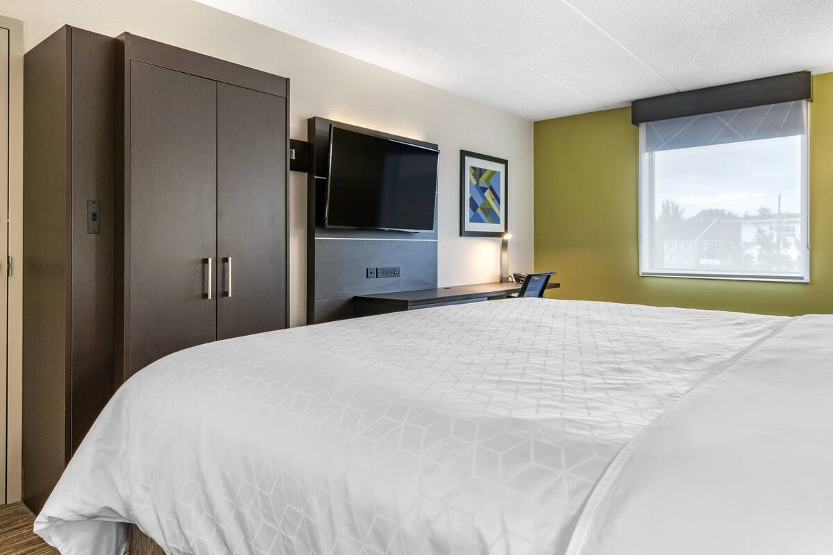 A hotel room with a large bed, a flat-screen TV, a desk, and a window with a view of buildings.