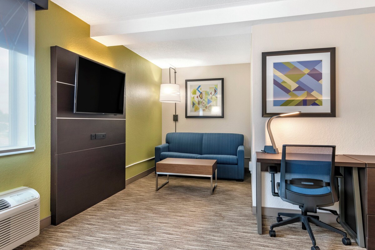A hotel room with a flat-screen TV, a sofa, a desk with a chair, and a lamp.