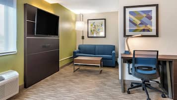 A hotel room with a flat-screen TV, a sofa, a desk with a chair, and a lamp.