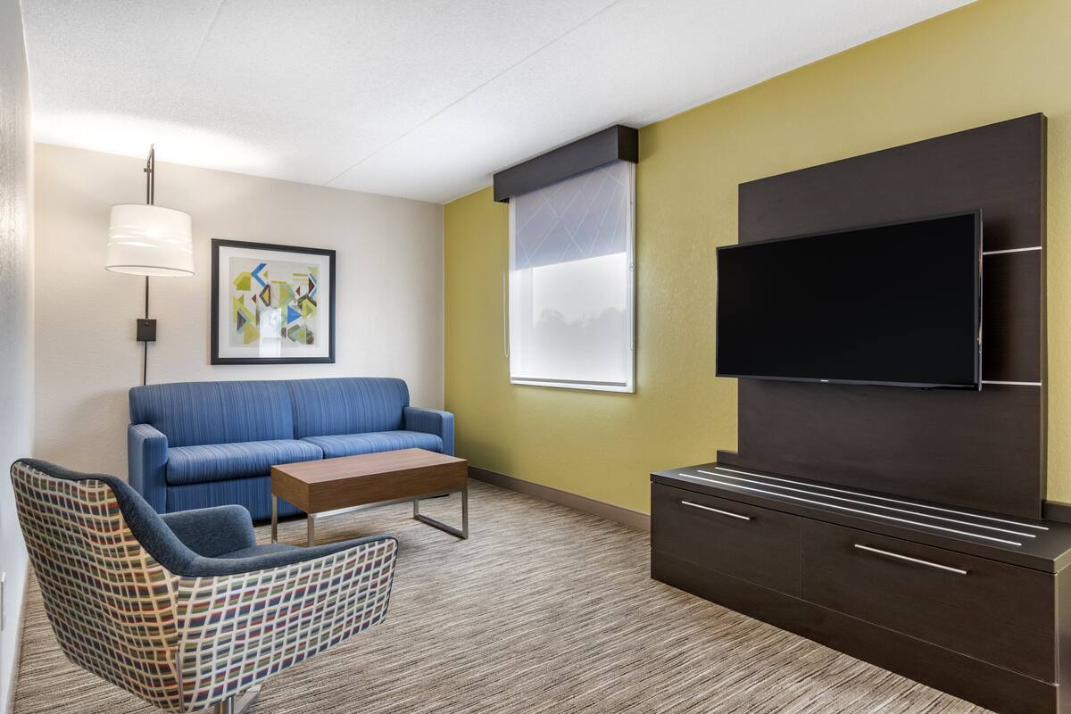 A modern living room with a blue sofa, a patterned armchair, a wooden coffee table, a flat-screen TV mounted on a dark cabinet, and a large window with blinds.