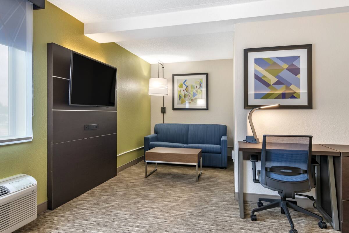 A hotel room with a flat-screen TV, a blue sofa, a wooden coffee table, a desk with a chair, and a framed abstract painting.