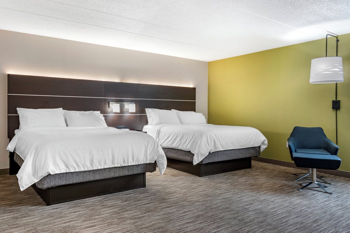 A hotel room with two beds, a nightstand, a lamp, and a chair.