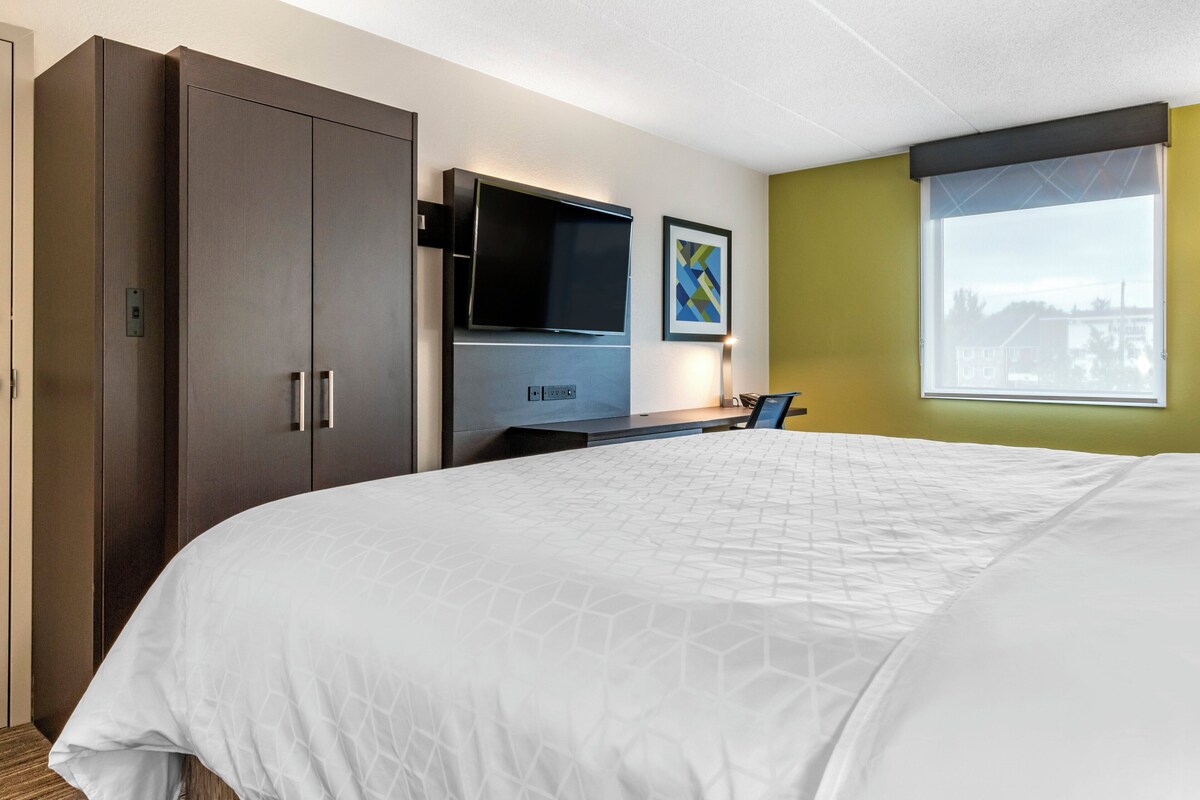 A hotel room with a large bed, a flat-screen TV, a desk with a chair, and a window with a view.