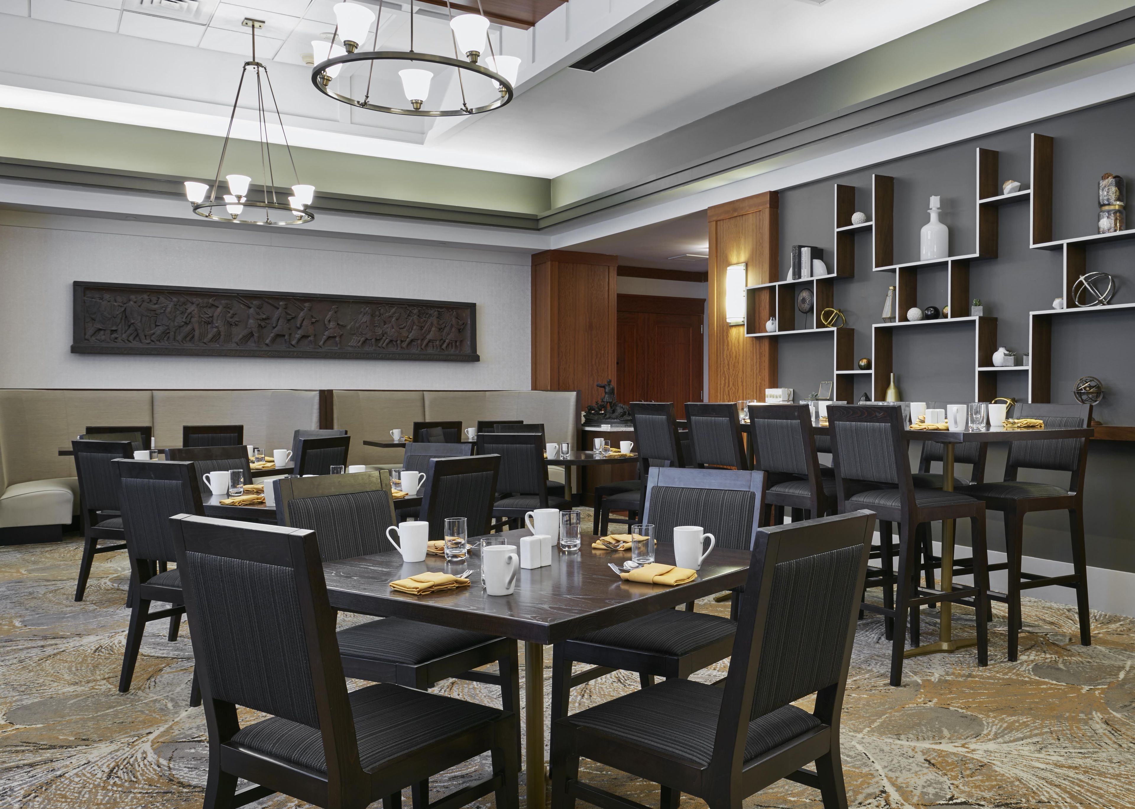 A hotel dining area with multiple tables and chairs, a chandelier, and wall-mounted shelves with decorative items.