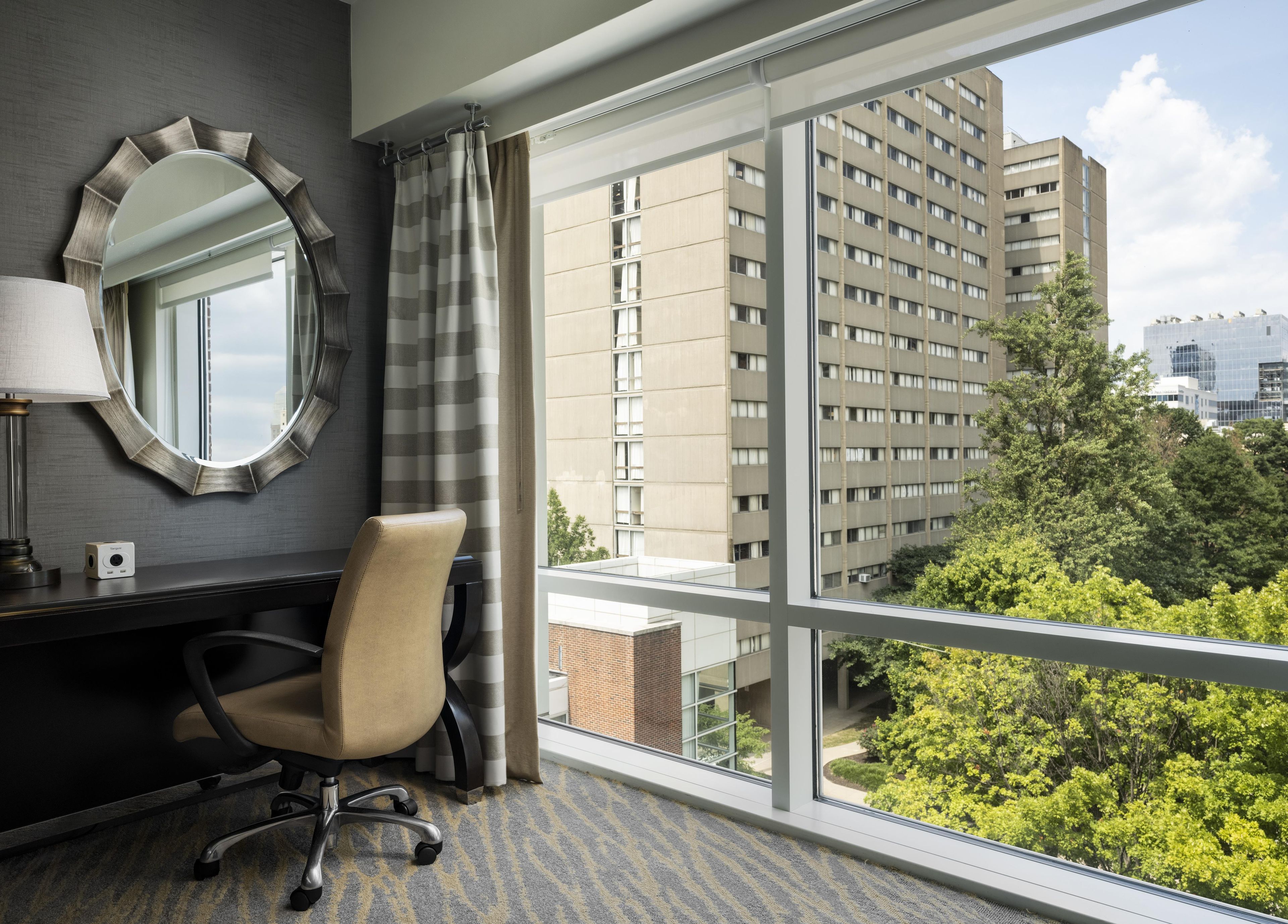 Ein Hotelzimmer mit großem Fenster, Schreibtisch, Stuhl, rundem Spiegel und Lampe.
