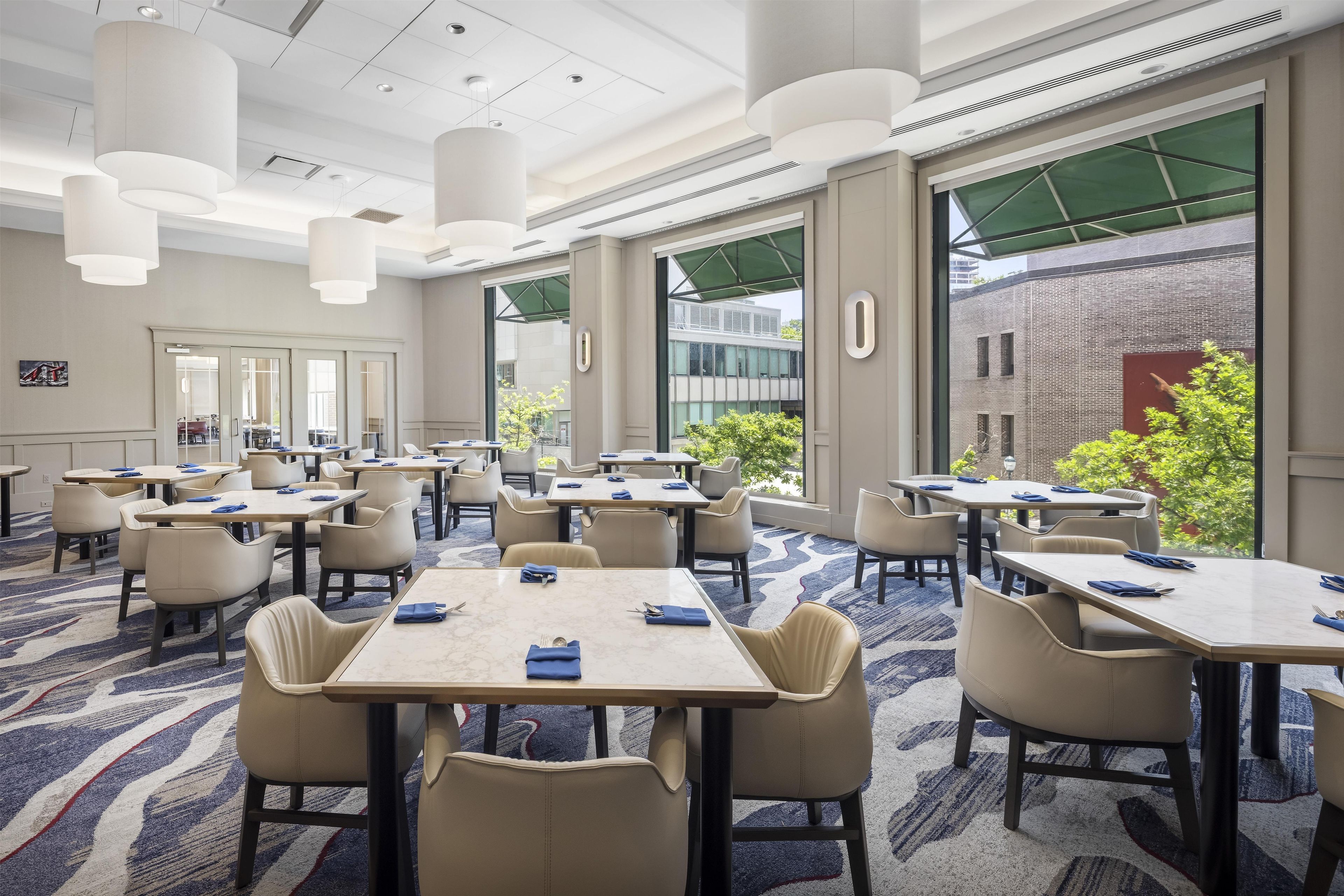 A hotel conference room with multiple tables and chairs, large windows, and a view of the outside.