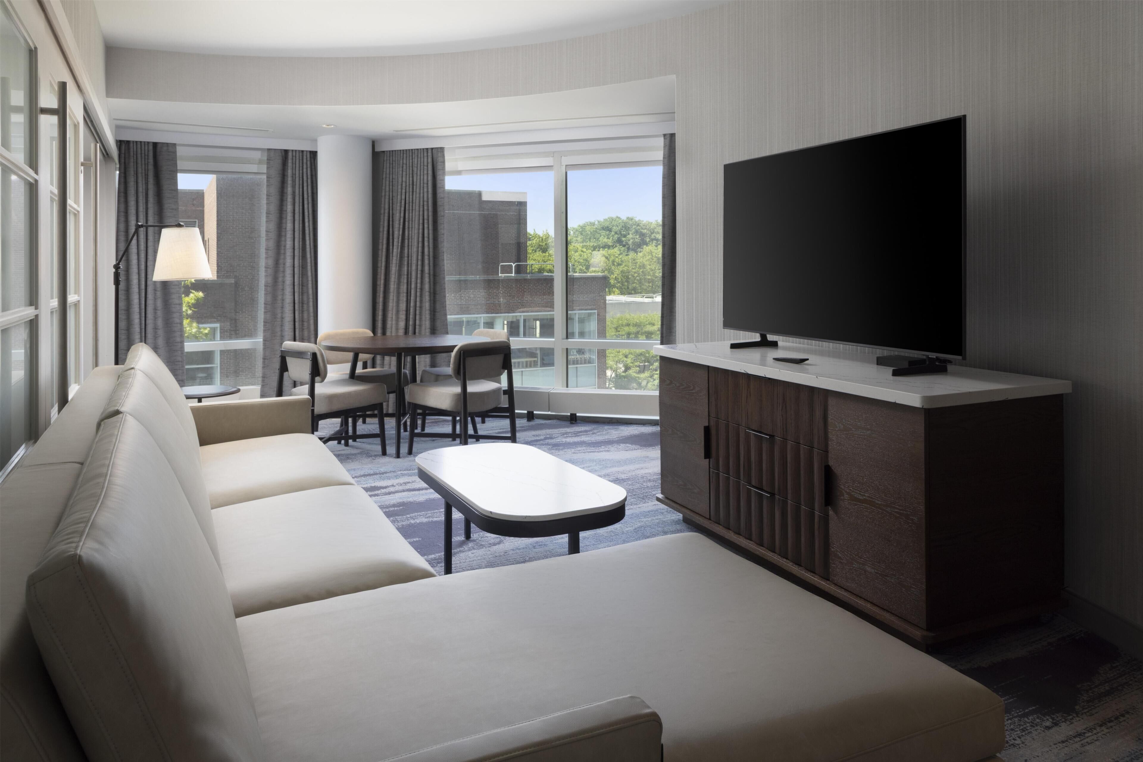 A modern hotel room with a sofa, a flat-screen TV, a dining table with chairs, and a balcony with a view of the city.