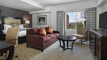 Quarto de hotel com cama, sofá, escrivaninha, cadeira, televisão e janela com vista para edifícios.