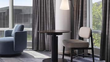Une chambre d’hôtel avec une table ronde, deux chaises, une lampe et de grandes fenêtres avec des rideaux.
