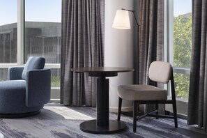 Une chambre d’hôtel avec une table ronde, deux chaises, une lampe et de grandes fenêtres avec des rideaux.