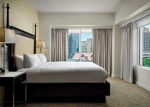 A hotel room with a large bed, a bedside table with a lamp, a view of the cityscape through floor-to-ceiling windows, and a carpeted floor.