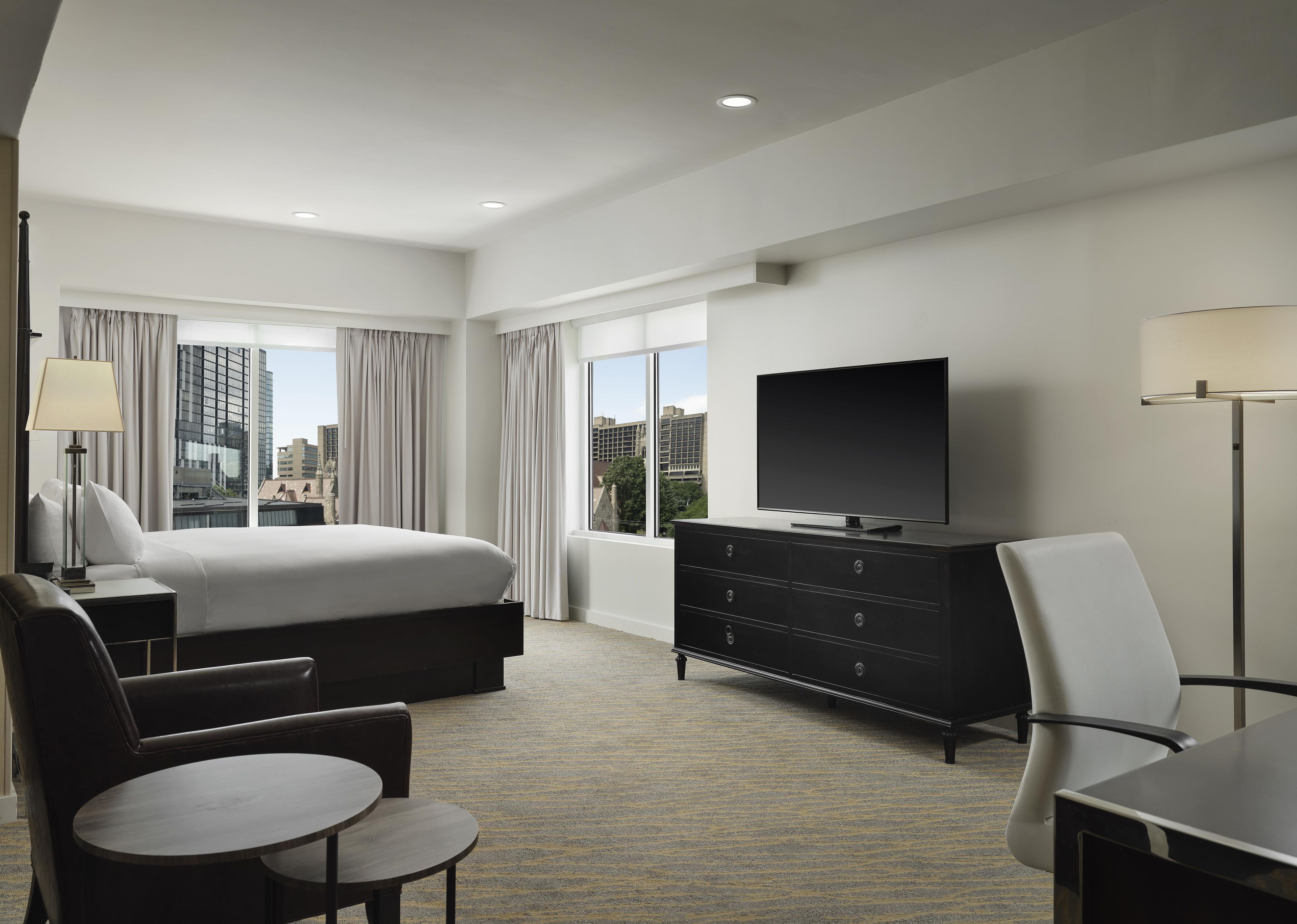 A hotel room with a bed, a flat-screen TV, a desk, a chair, and a view of the cityscape through large windows.