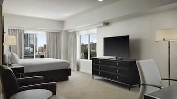 A hotel room with a bed, a flat-screen TV, a desk, a chair, and a view of the cityscape through large windows.