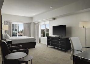 A hotel room with a bed, a flat-screen TV, a desk, a chair, and a view of the cityscape through large windows. - The Inn at Penn, A Hilton Hotel (Philadelphia)