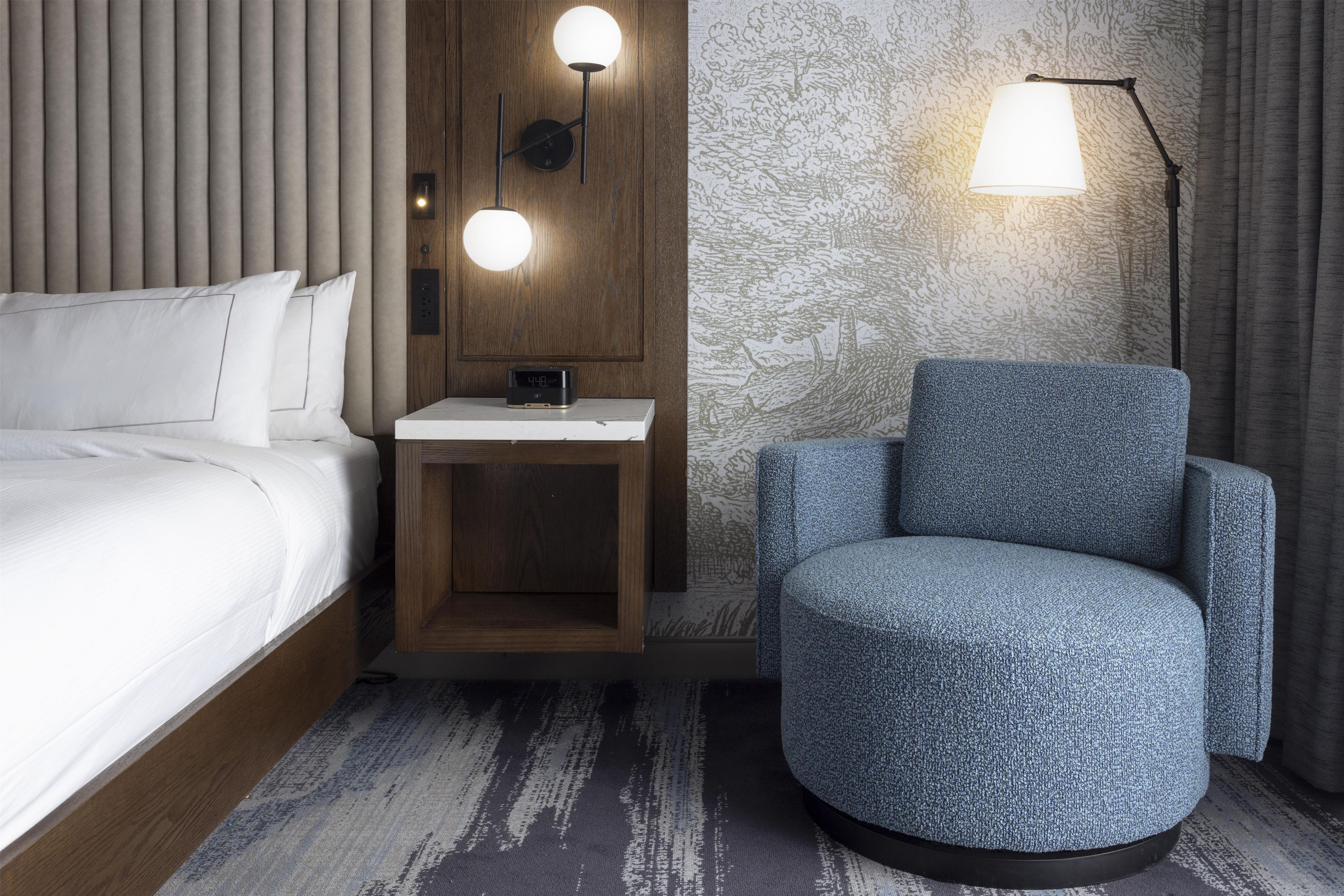 Une chambre d’hôtel avec un lit, une table de chevet avec une horloge, une lampe et un fauteuil bleu.