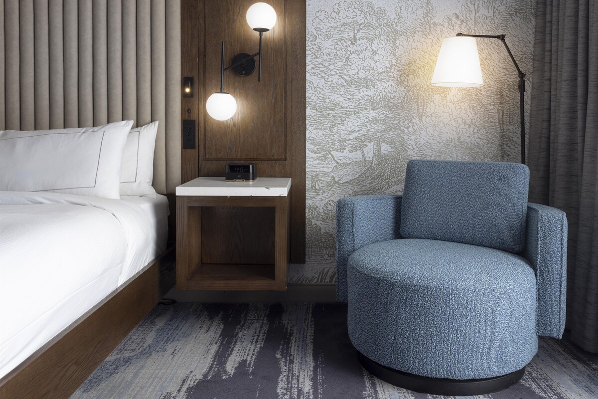 A hotel room with a bed, a nightstand with a clock, a lamp, and a blue armchair.