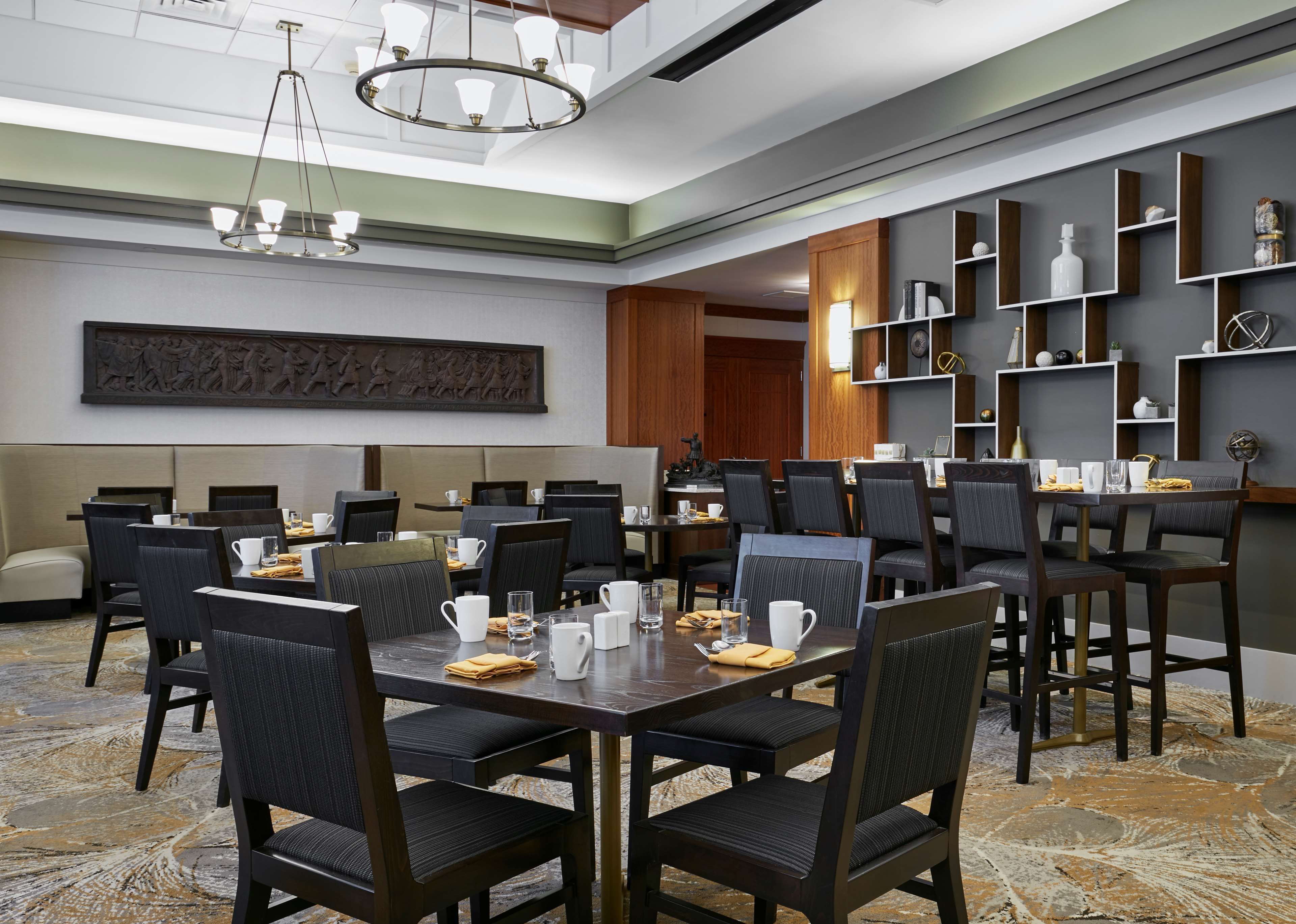 A hotel dining area with multiple tables and chairs, a chandelier, and wall-mounted shelves with decorative items.