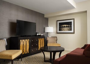 Habitación de hotel con televisor de pantalla plana, aparador con cajones, mesa redonda, dos sillones, sofá, lámpara y un cuadro en la pared.