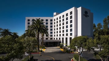 A modern hotel building with a flat roof, multiple windows, and a parking lot with trees and shrubs.