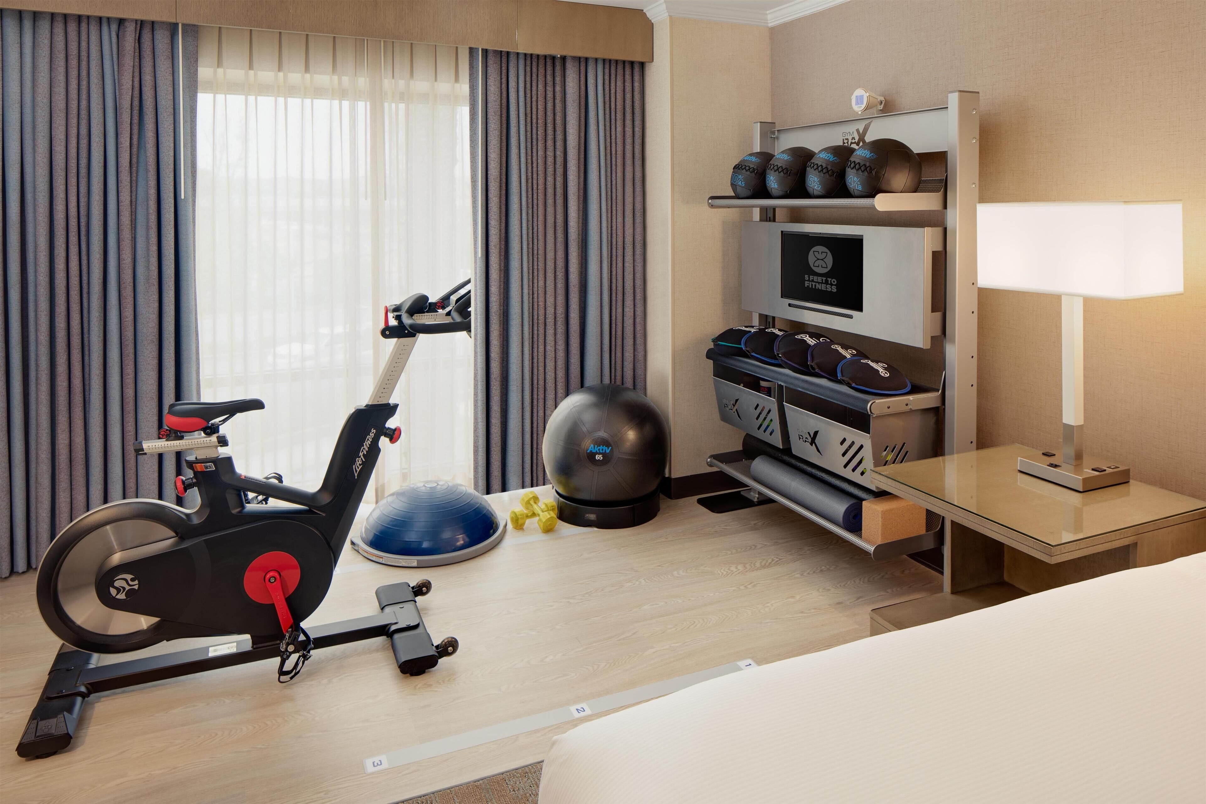 A hotel room with a bed, a desk, a treadmill, a set of dumbbells, a kettlebell, and a television.