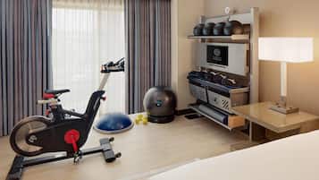 A hotel room with a bed, a desk, a treadmill, a set of dumbbells, a kettlebell, and a television.