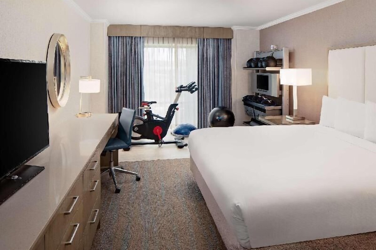 Een hotelkamer met een bed, bureau, stoel, een hometrainer, televisie en een grote spiegel.