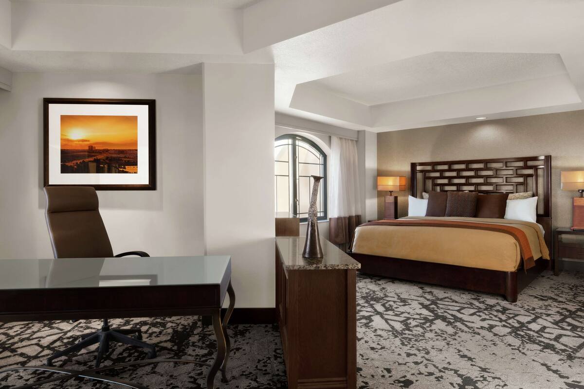 Een hotelkamer met een bed, een bureau, een stoel, een ingelijst schilderij aan de muur en een raam met gordijnen.