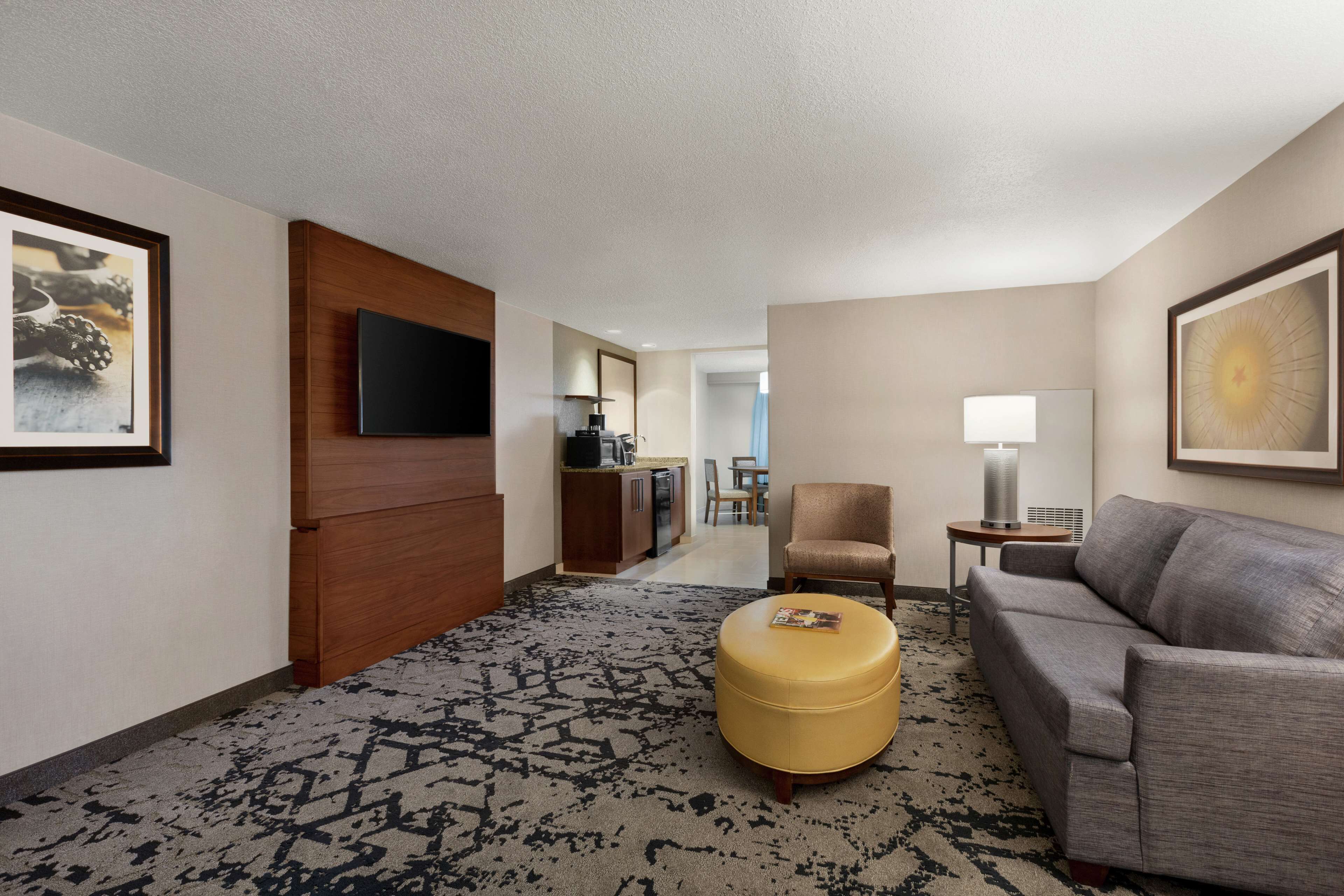 A hotel room with a grey sofa, a yellow ottoman, a TV mounted on a wooden panel, a dining area in the background, and a framed picture on the wall.
