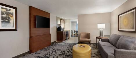 A hotel room with a grey sofa, a yellow ottoman, a TV mounted on a wooden panel, a dining area in the background, and a framed picture on the wall.