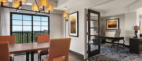 A hotel room with a dining area, a desk, and a view through a large window.