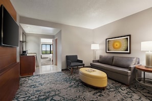 A modern living room with a flat-screen TV, a sofa, a chair, a coffee table, and a lamp. - Embassy Suites by Hilton Dallas DFW Airport North (Grapevine)