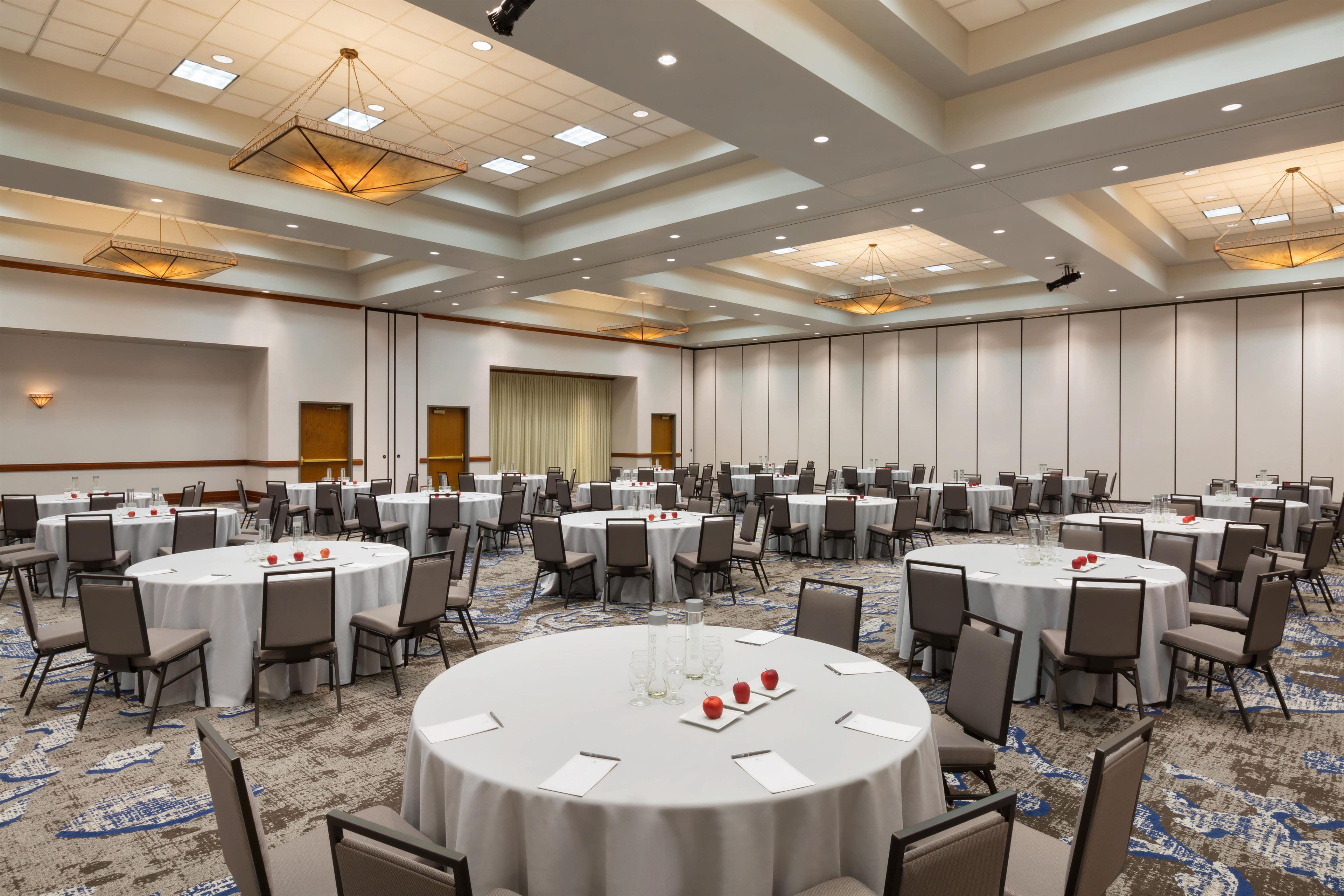Une grande salle de conférence dotée de nombreuses tables rondes, chacune étant agrémentée de chaises et de couverts, ainsi que de verres à eau et d'assiettes contenant des pommes.
