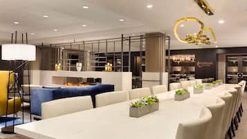 A modern hotel dining area with a long table, chairs, and a central seating area with a blue sofa and a fire feature.