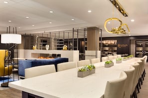 Un espace de restauration moderne dans un hôtel, avec une longue table, des chaises et un coin salon central agrémenté d’un canapé bleu et d’un foyer.