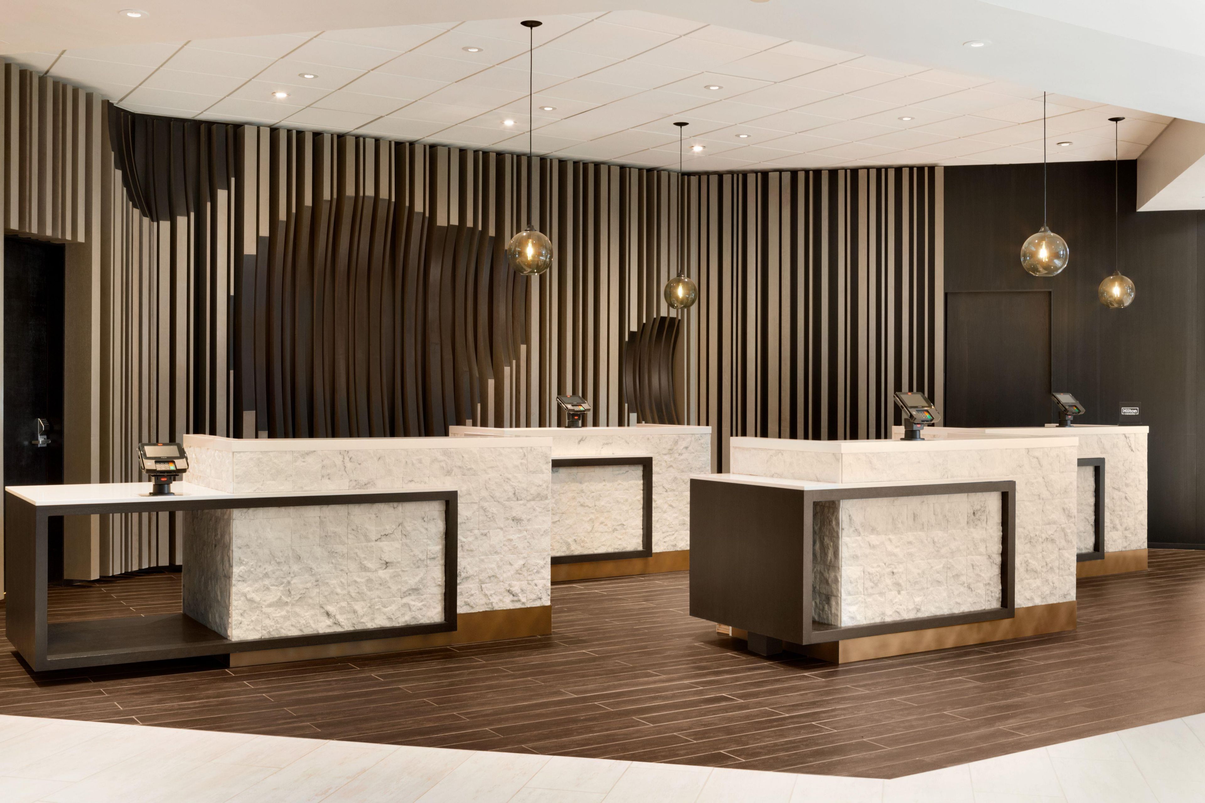 Zona de recepción de hotel con mostradores de mármol, diseño moderno con listones verticales de madera e iluminación colgante.