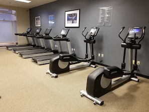 A row of treadmills in a fitness center with a grey wall, a projector screen, and a sign listing fitness center channels.