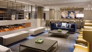 Un hall d’hôtel moderne avec un plafond haut, une cheminée, des espaces de détente et un grand tableau abstrait.