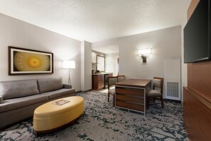 A hotel room with a sofa, ottoman, dining table, chairs, a flat-screen TV, a painting, a lamp, and a kitchenette.