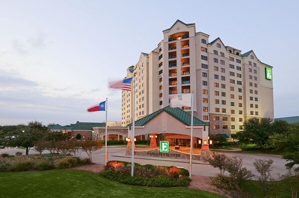 A multi-story hotel with a prominent 'E' sign, a flagpole with the American flag, and a well-maintained landscaped entrance. - Embassy Suites by Hilton Dallas DFW Airport North (Grapevine)
