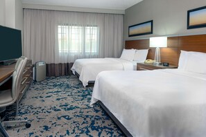 Ein Hotelzimmer mit zwei Betten, einem Schreibtisch, einem Stuhl, einem Fernseher und einem Fenster mit Vorhängen.