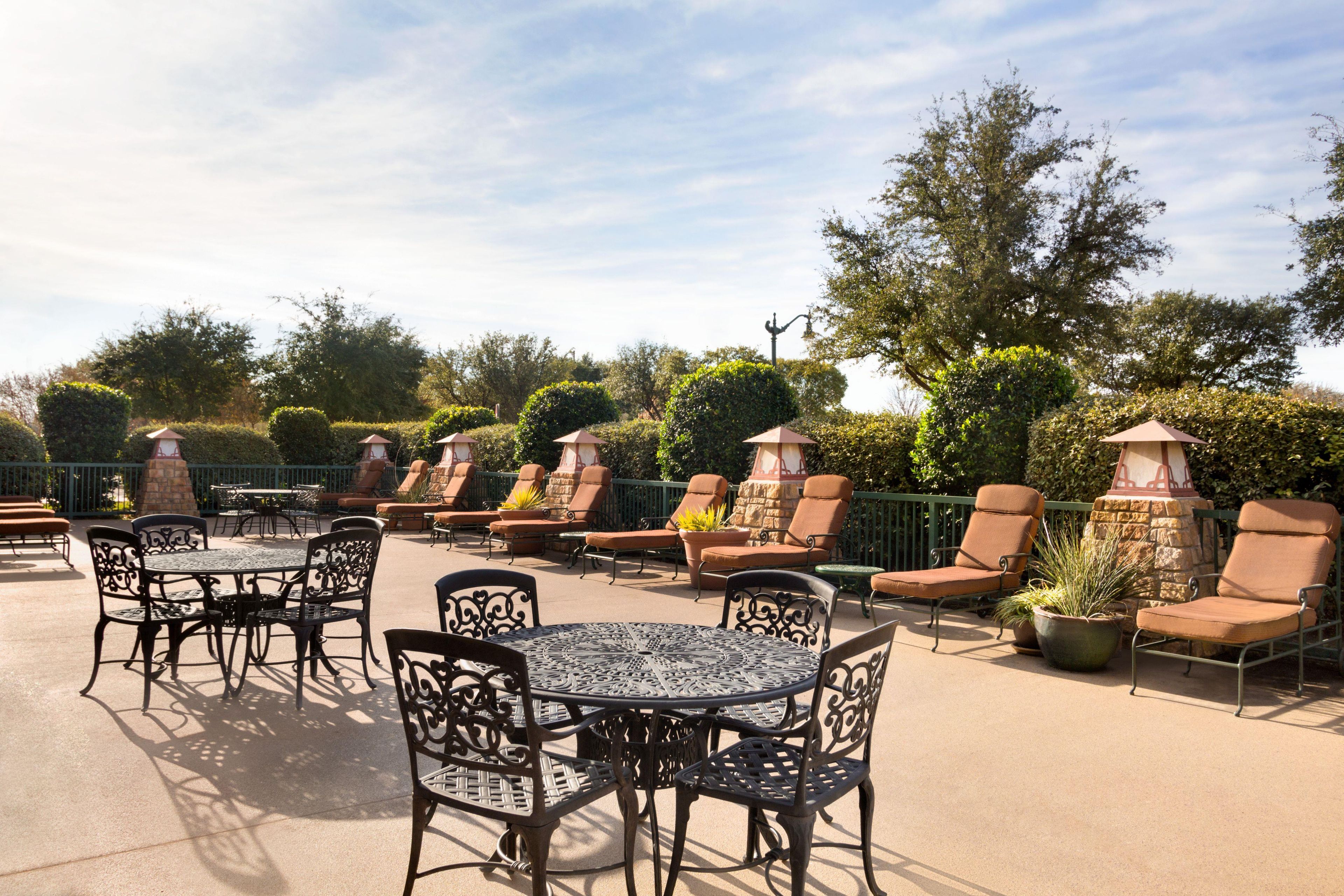 An outdoor patio area with tables and chairs, surrounded by greenery and a clear sky.