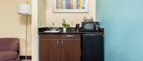 A compact kitchenette with a microwave, a small refrigerator, a sink, and a cabinet. There is a floor lamp and a potted plant on the counter.