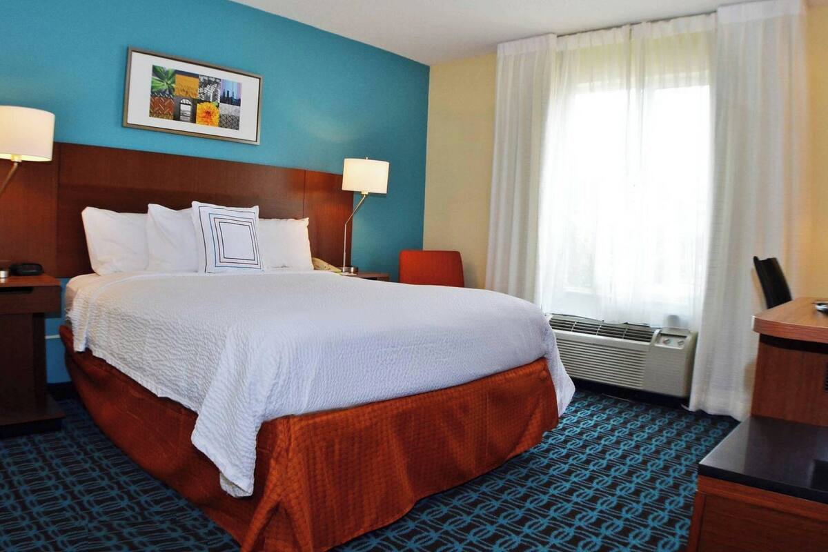 A hotel room with a large bed, a desk, a chair, a nightstand, a lamp, and a window with curtains.