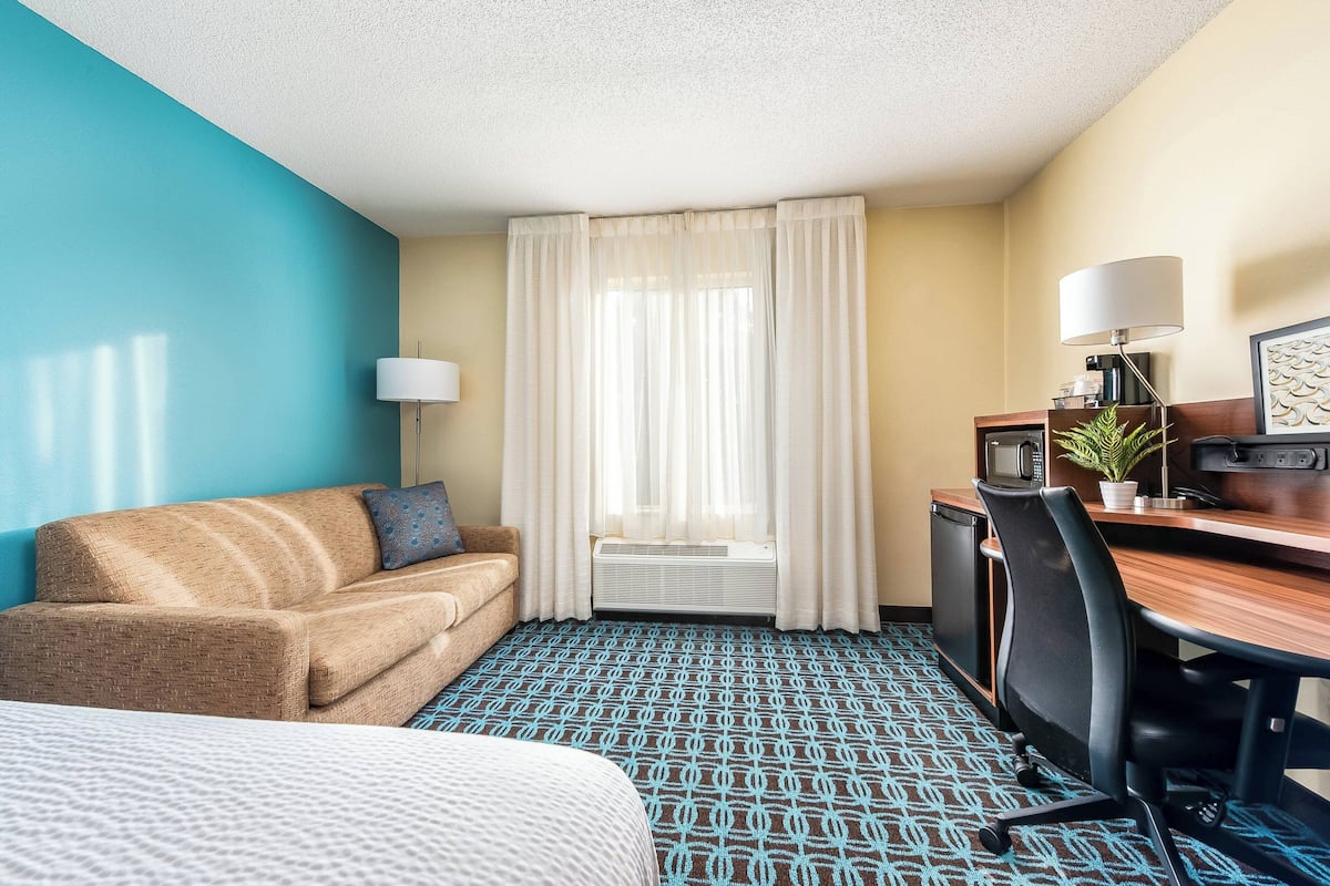 A hotel room with a bed, a sofa, a desk with a computer, a lamp, and a window with curtains.