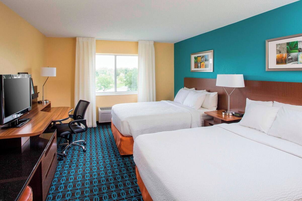 A hotel room with two beds, a desk with a television, a chair, and a window with curtains.