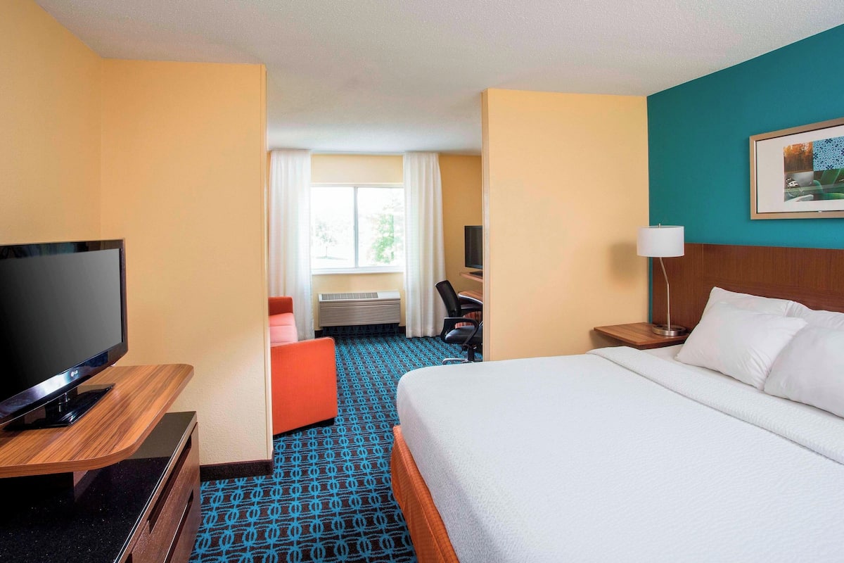 A hotel room with a bed, a TV on a wooden stand, a desk with a chair, and a window with curtains.