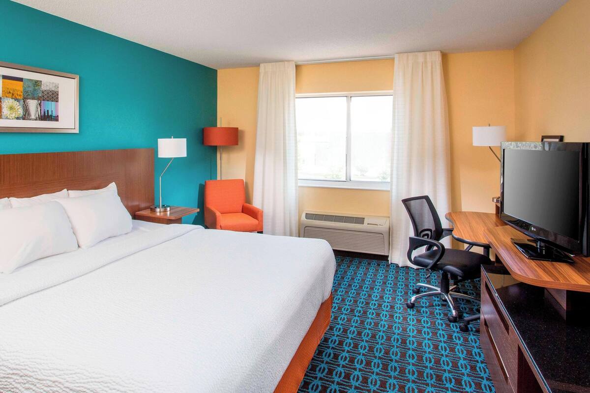 A hotel room with a large bed, a desk with a TV, a chair, and a window with curtains.