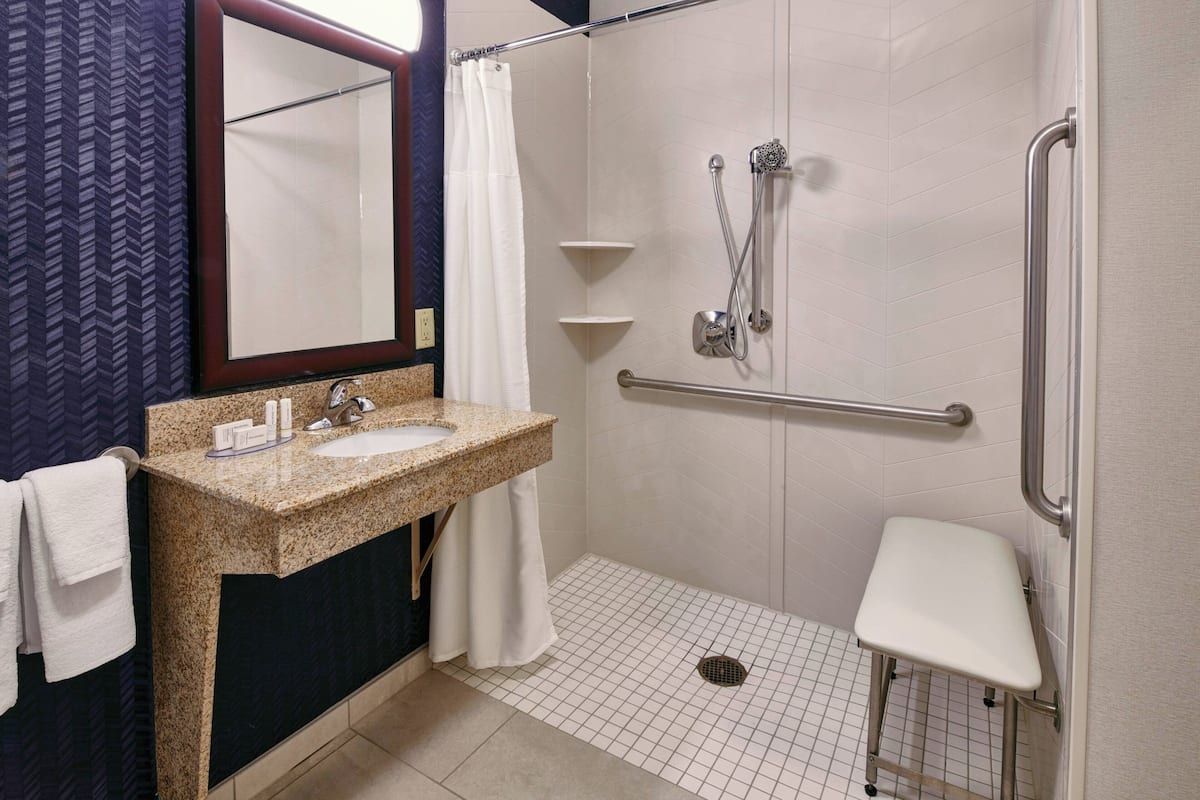 A bathroom with a granite sink, white tiles, a shower with a handheld showerhead, a grab bar, and a white bench.