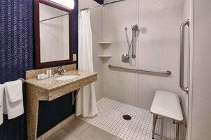 Un baño con lavamanos de granito, azulejos blancos, ducha con cabezal de ducha manual, barra de apoyo y banco blanco.