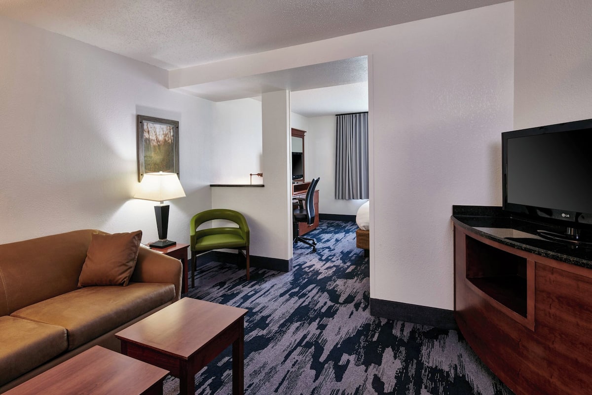 A hotel room with a brown leather sofa, a wooden coffee table, a TV on a wooden stand, a green chair, a desk with a lamp, and a carpeted floor.